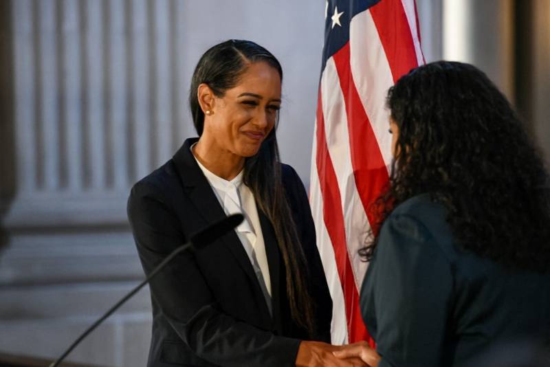 A woman in a black blazer shakes hands with Mayor London Breed, while the two stand in front of an American flag.
