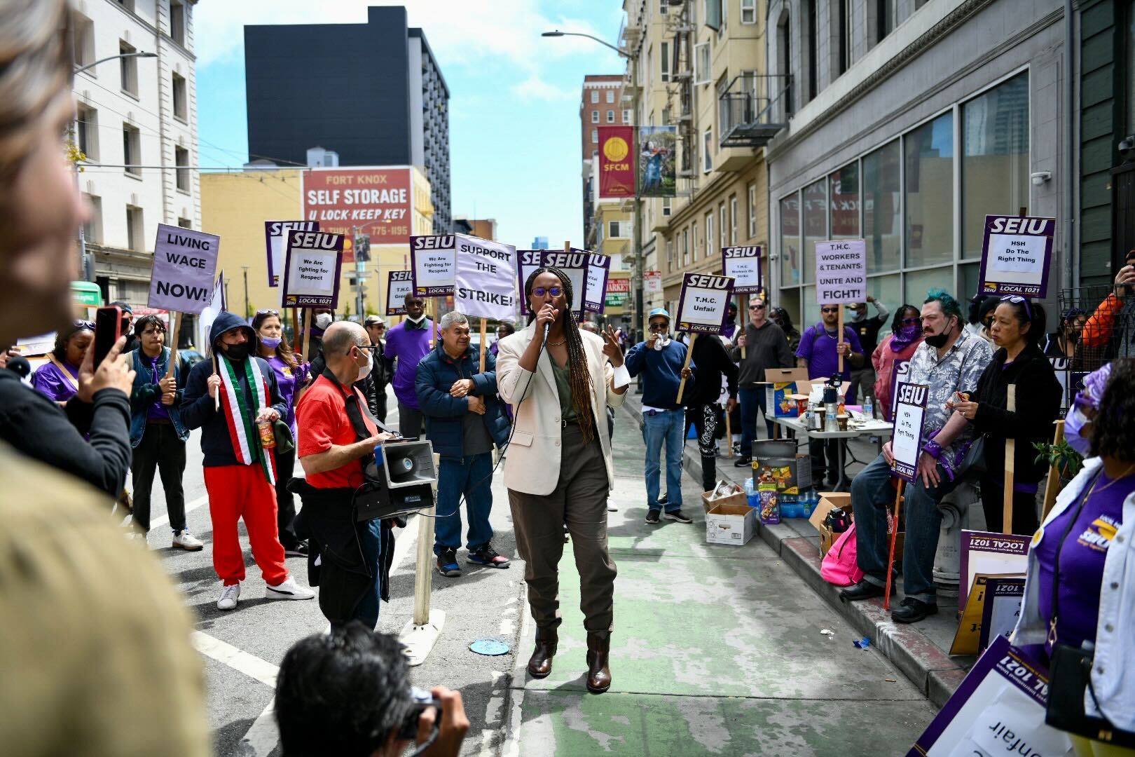 A woman speaking into a microphone stands on the street, surrounded by protesters holding signs.