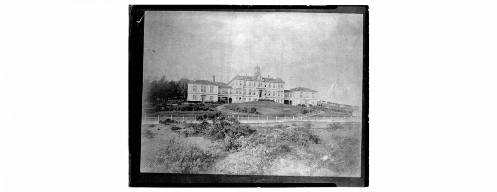 An old black and white photo of a large institutional looking building on a hill with open fields around it