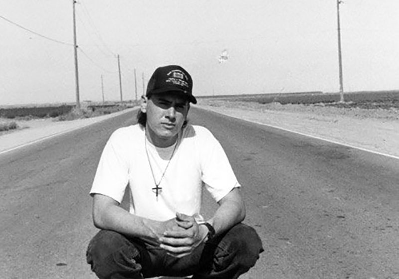A black and white photo of a young man in a white shirt and black cap crouches on an empty roadway