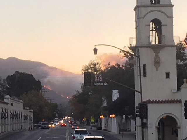 Thick golden yellow smoke clogs the sky over a dark mountain in the background, where a line of fire is visible in the distance. In the middle distance and foreground is the main street of a town, framed on the left and right by dark green trees and white buildings. On the right is a tall building with a tower. On the left are low buildings. The middle of the picture shows cars moving in both directions with headlights on.