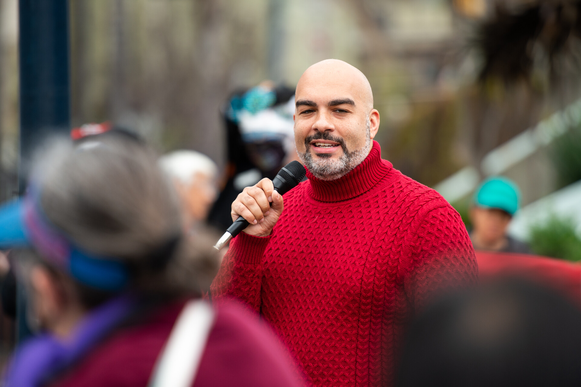 A bald man with a salt and pepper beard and red sweater holds a microphone whiel standing among a crowd, outside. 