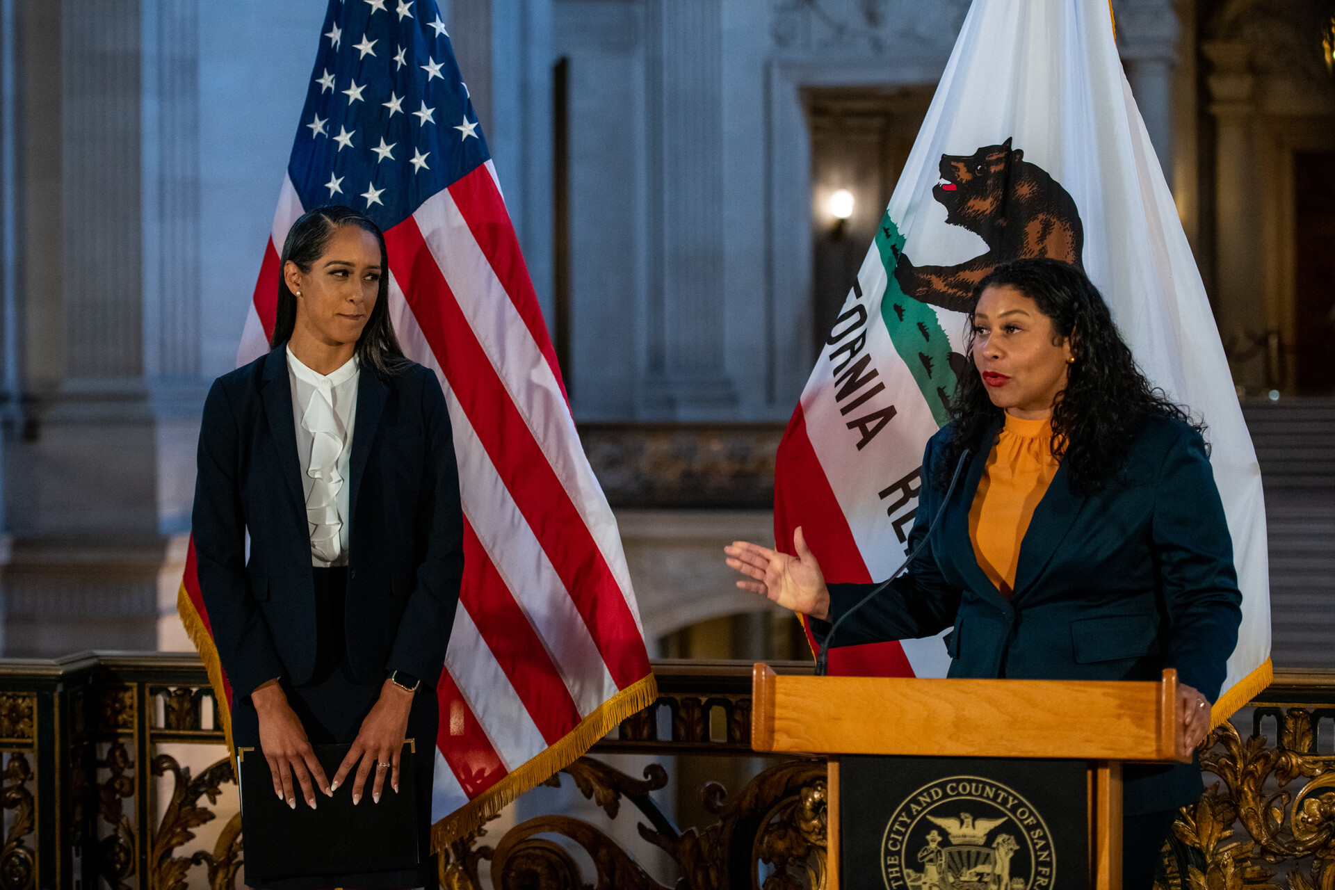 Mayor London Breed stands at a podium, her right hand extended,with a California and American flag behind her, introducing Brooke Jenkins - standing to her right — as San Francisco's new district attorney.