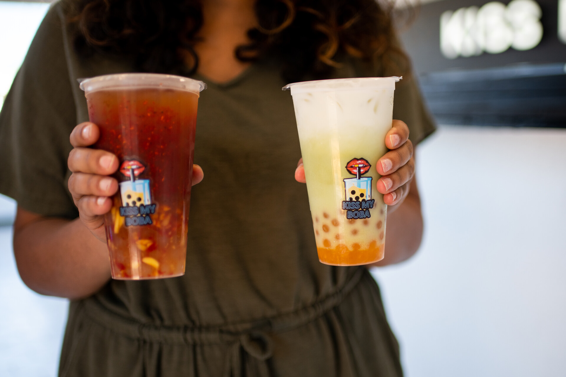 detail photo of two hands holding large plastic cups, one filled with a brownish reddish drink, the other a white and orange drink with boba at the bottom