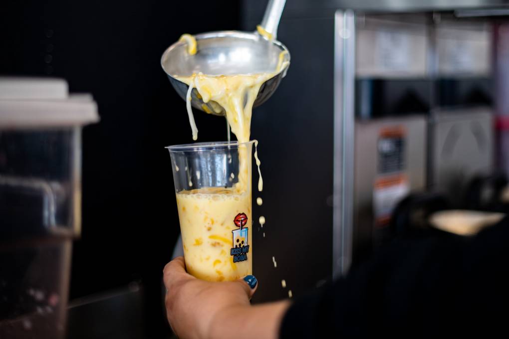hand holds cup while other hand ladles creamy white and orange drink, with visible chunks of mango and shredded coconut