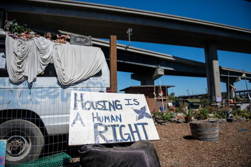 A sign says, 'Housing is a Human Right' at an encampment under a highway overpass.