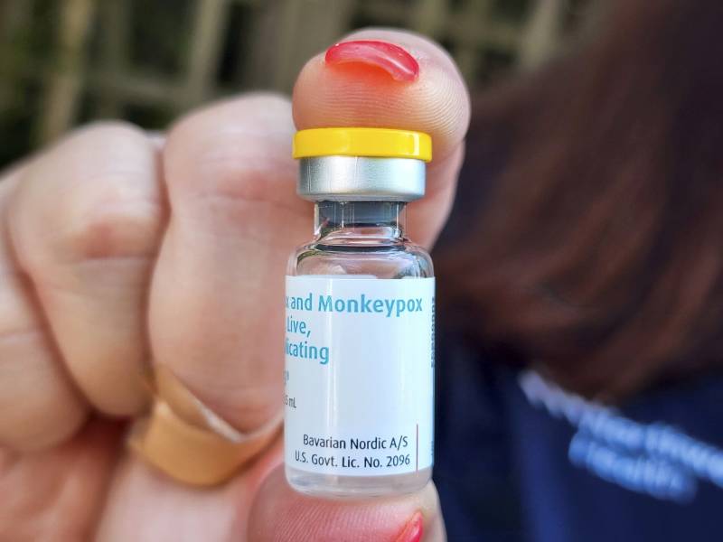 A right hand with nails painted red and a bandaid visible on the tip of the third finger holds a vial of the monkeypox vaccine Jynneos. The cap of the vial is bright yellow, and the label is white with blue lettering.