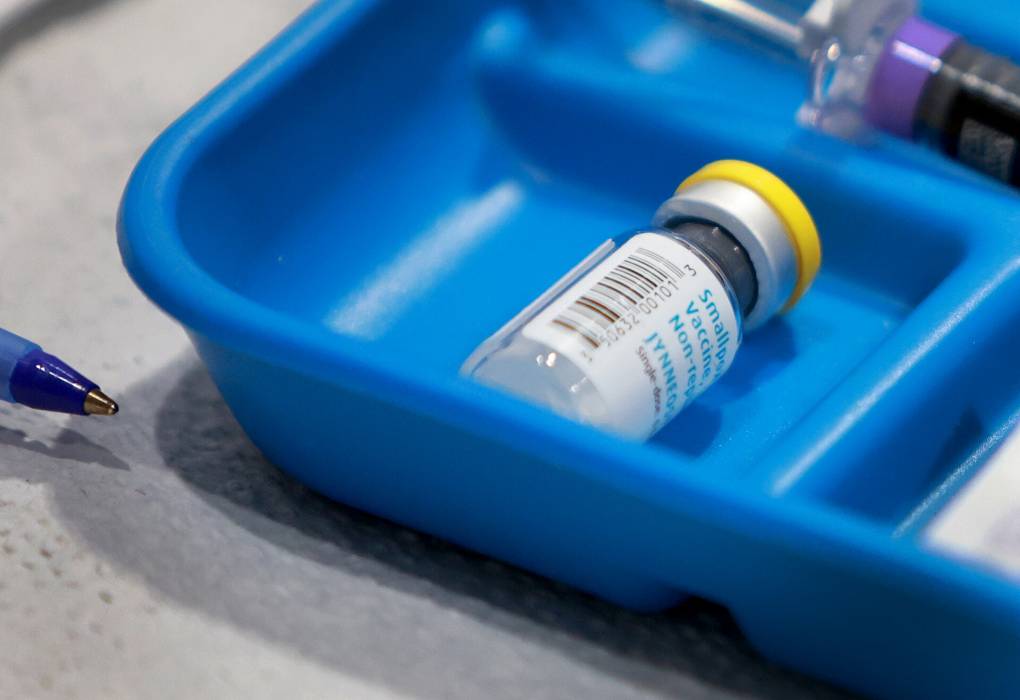 A vial of the Jynneos vaccine lies on its side in a shallow bright blue container, with a needle seen behind it out of focus.
