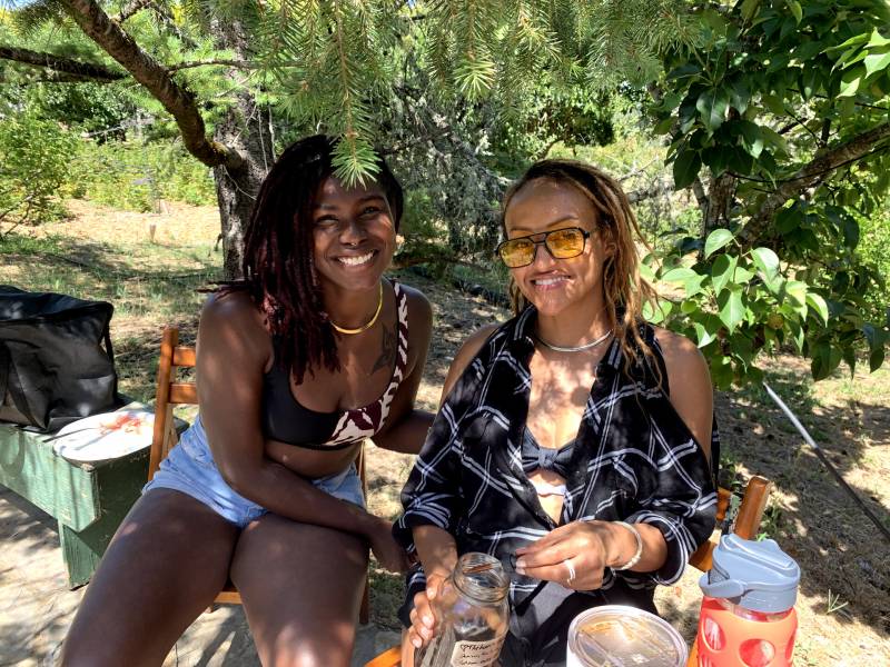 two smiling Black women sit under a tree on an orchard