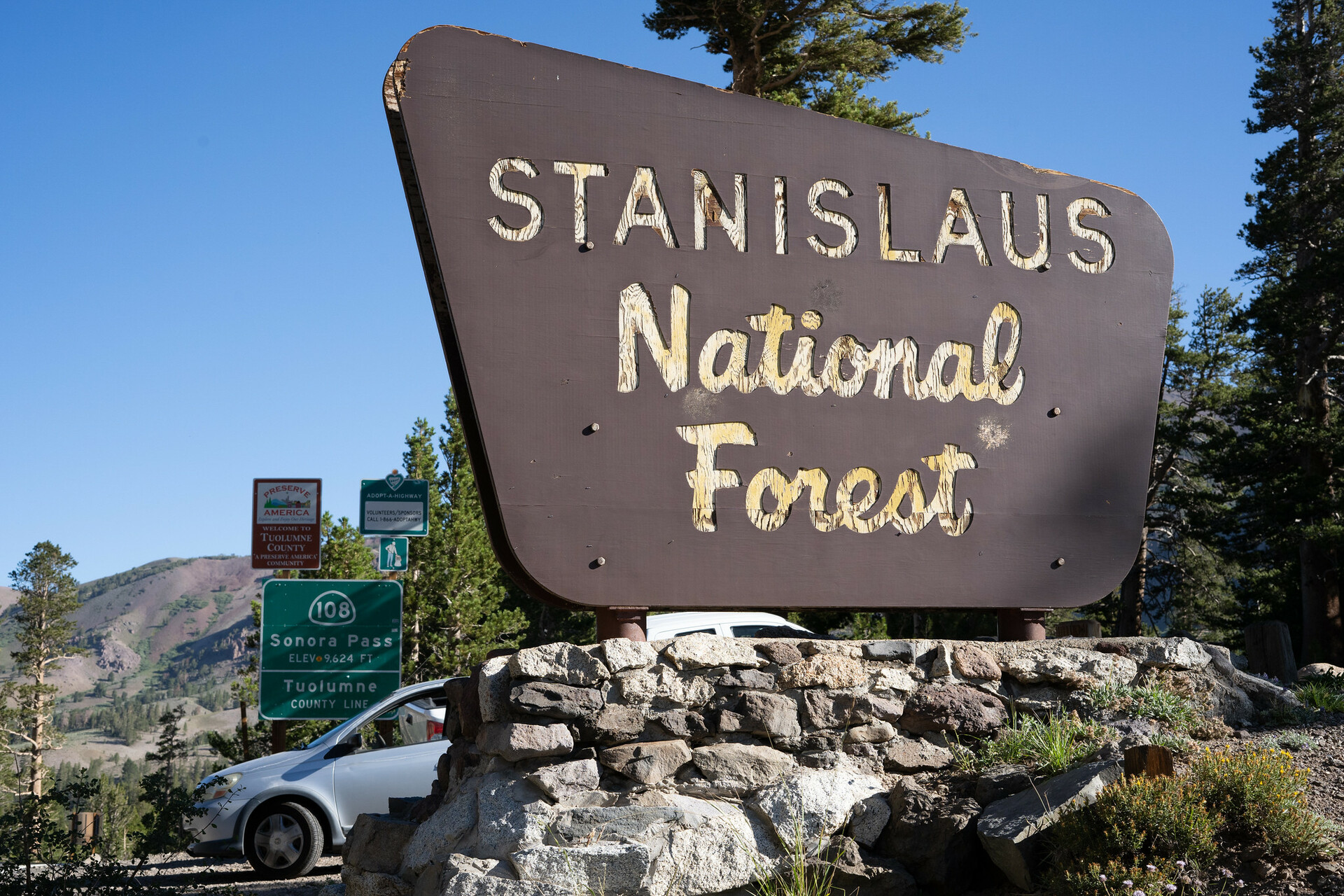 A large brown wooden sign that says STANISLAUS NATIONAL FOREST in yellow lettering, with a silver car visible behind it and a hill in the background