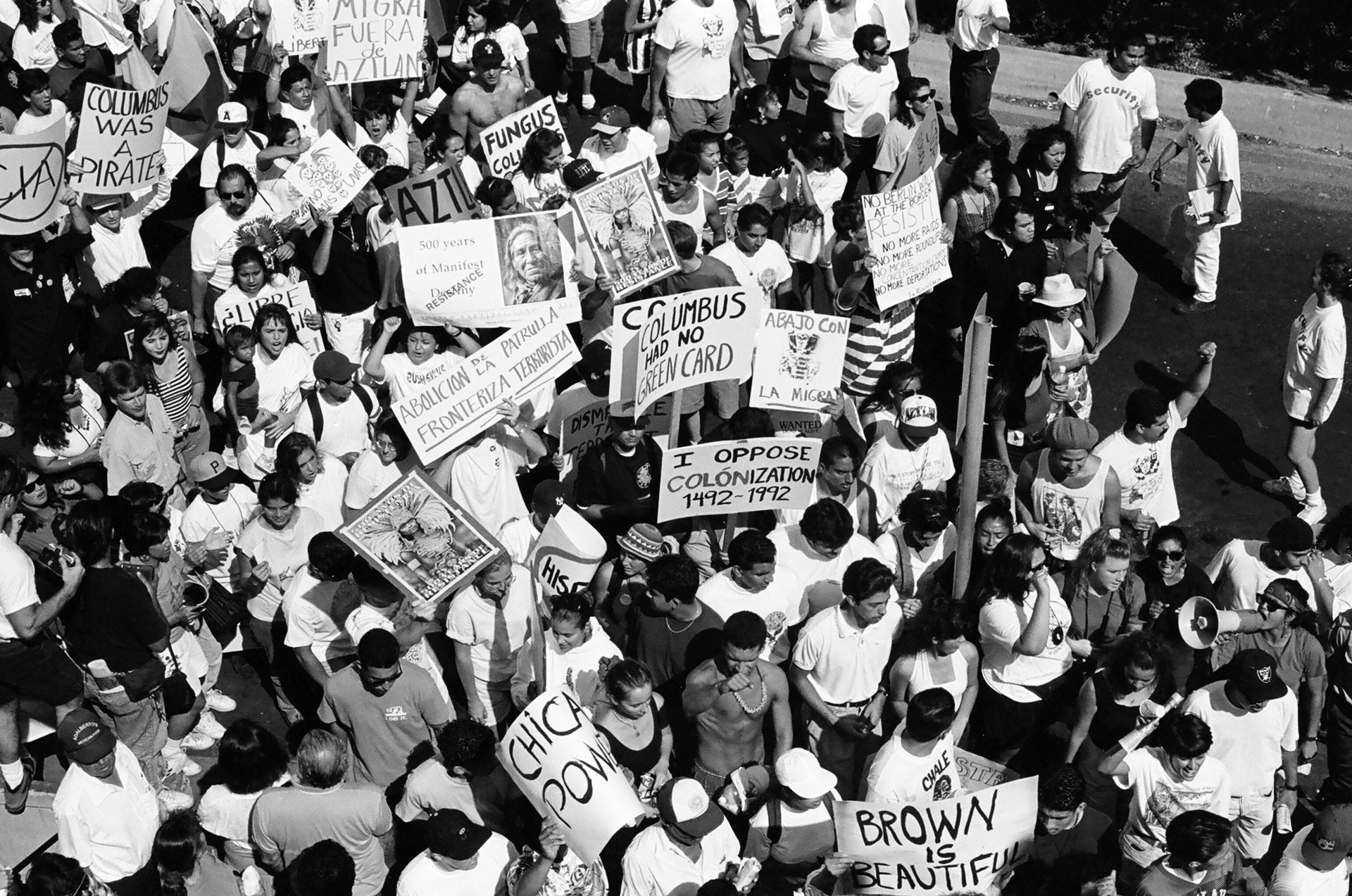aerial black and white photo of young activists holding signs reading 'Columbus had no green card' and 'Chicano power' and 'brown is beautiful'