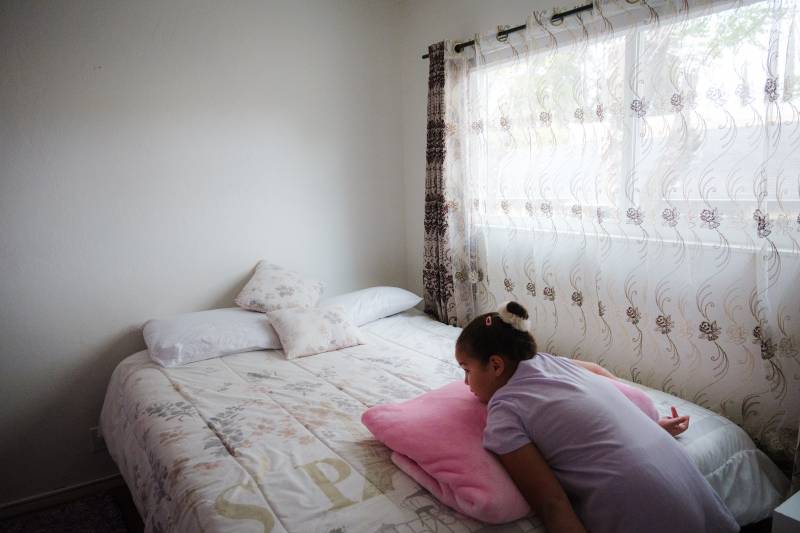 A young girl in a lavender cotton shift with short sleeves stands at the end of a bed, leaning over and resting her torso on top of a folded pink blanket. Her brown hair is pulled back into a bun, with a fuzzy white band around it, and a pink clip on the side of her head. Light filters through a wide window covered with translucent white curtains that are patterned with rows of pink and grey roses and swirls. The quilt on her bed is white with pale flowers in pink and lavender. Two throw pillows in the same fabric are tossed against two white bed pillows at the head of the bed. The walls of the room are white. 