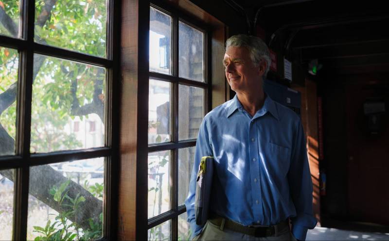 A man with rosy skin and silve-and-grey hair stands in a window looking out at the green leaves of a tree. Sunlight dapples his blue shirt. He wears slate green khaki pants and holds a grey zipped computer case under one arm. The window frames are dark wood.