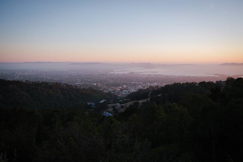 A view from high in the forested hills over the Bay Area. In the foreground are rolling hills covered with oak and pine, with a few mansions visible in nooks and crannies of the canyons. In the middle of the photo is a wide urban spread o, with the bay in the distance on the right of the image, and the Bay Bridge a faint line across. Above the urban landscape are bluish hills in the distance and a line of orange and yellow light on the horizon as the sun rises into a pale blue sky.