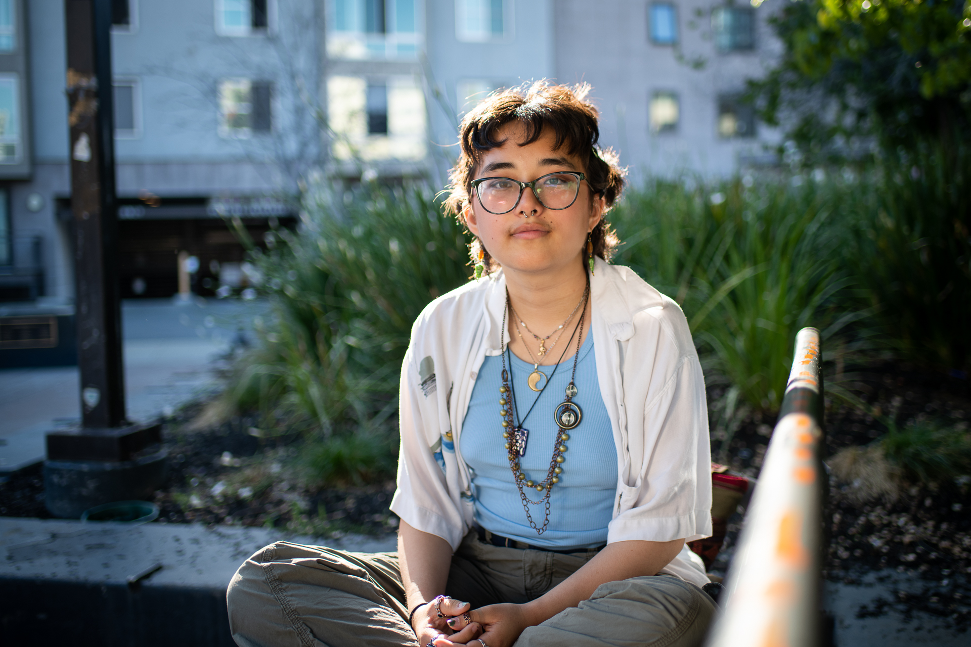 a young woman with short hair and glasses in a blue shirt sits cross-legged in a park