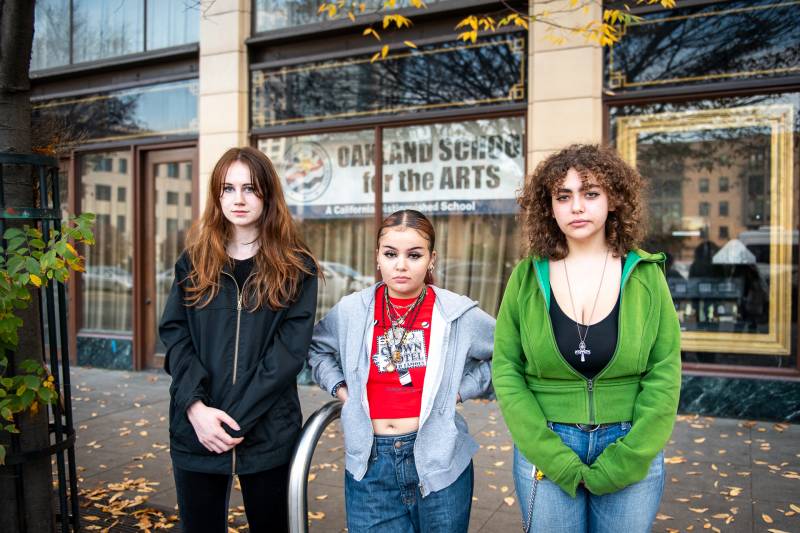 three young women stand looking sternly into the camera outside a school building