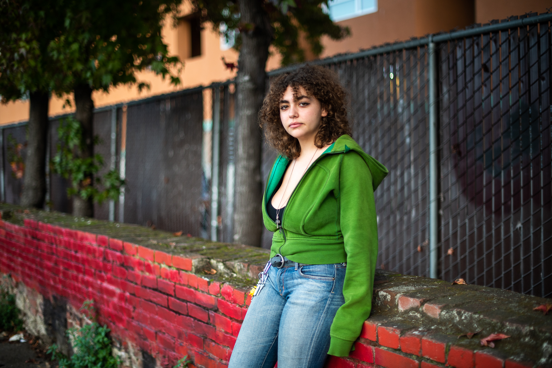 a young woman in jeans and a green hoodie leans against a brick wall
