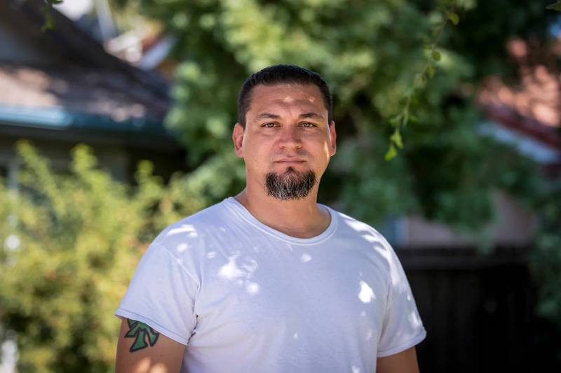 A young Latino man in a white t-shirt stands for a portrait in front of green trees