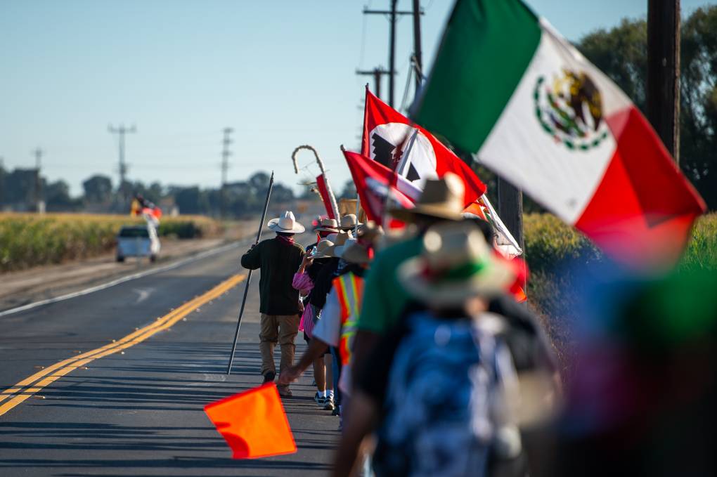 line of marchers carrying union flags and a mexican flag walk down a rural 2-lane highway in the hot sun