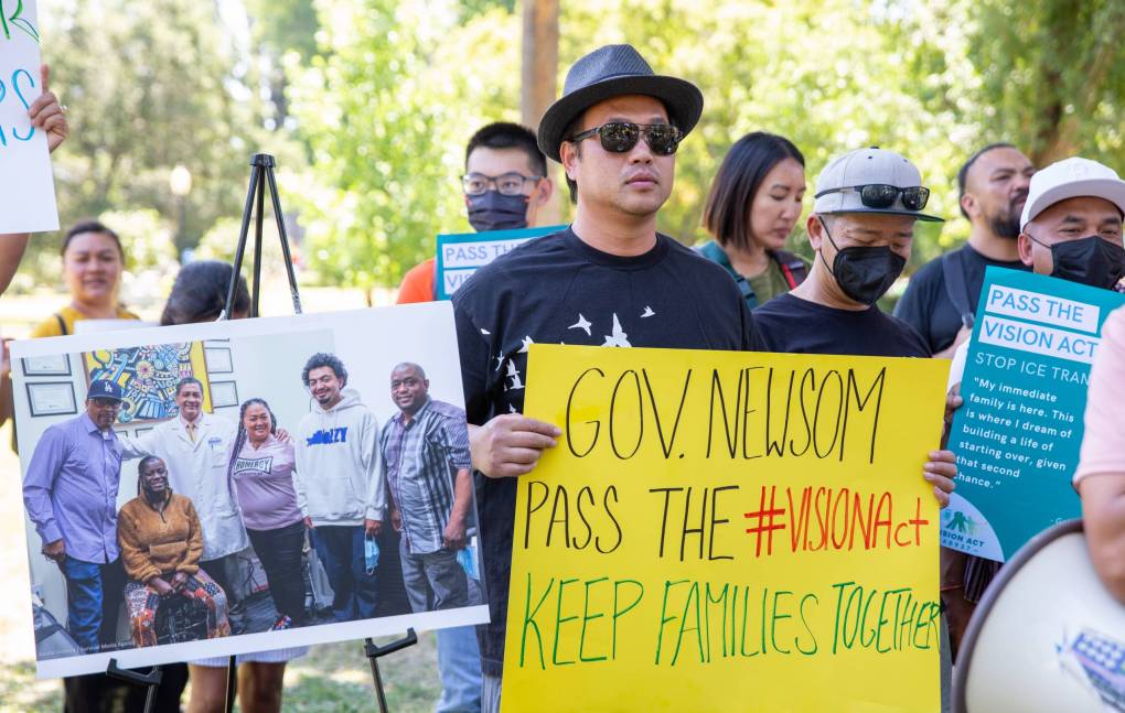 Demonstrators stand in a group, holding signs and photos. One sign says: 'Gov. Newsom Pass The #VisionAct Keep Families Together'