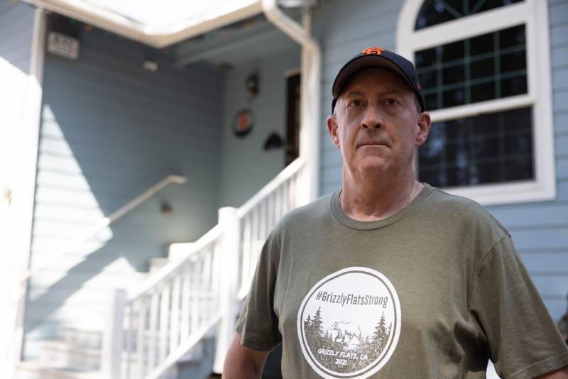 A man with pale skin wearing a light sage t-shirt and SF Giants ball cap stands in front of a light blue home with white painted wood railing and steps leading to the front door. A round logo on his shirt reads "hashtag Grizzly Flats Strong" over an image of trees and a mountain.