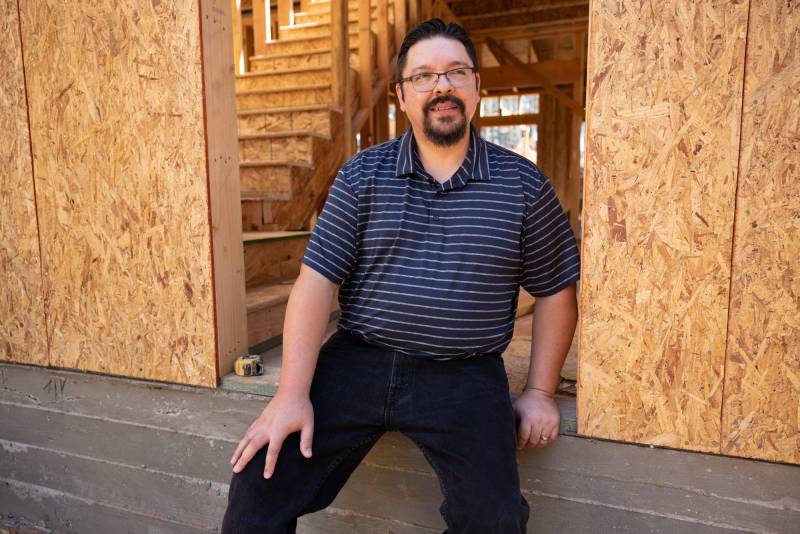 A man with light skin, black hair and a black beard sits on the foundation of a home being built. He's sitting in a doorway, framed walls rising beside him. He's wearing a blue short-sleeved shirt with narrow white stripes and blue jeans. 