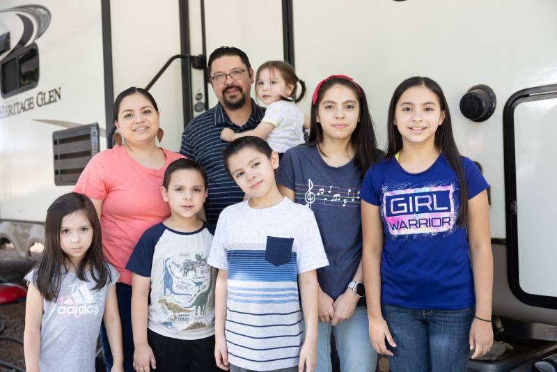 A man and his wife with their six children and in front of an RV where they live. The family has light brown skin and dark hair. The children range in age from 5 to 11. They wear jeans with t-shirts in blue, white, or pink.