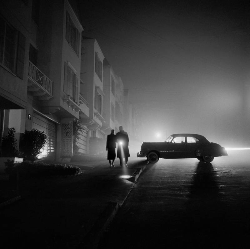 Black and white night-time image with man, woman and car silhouetted in fog. 