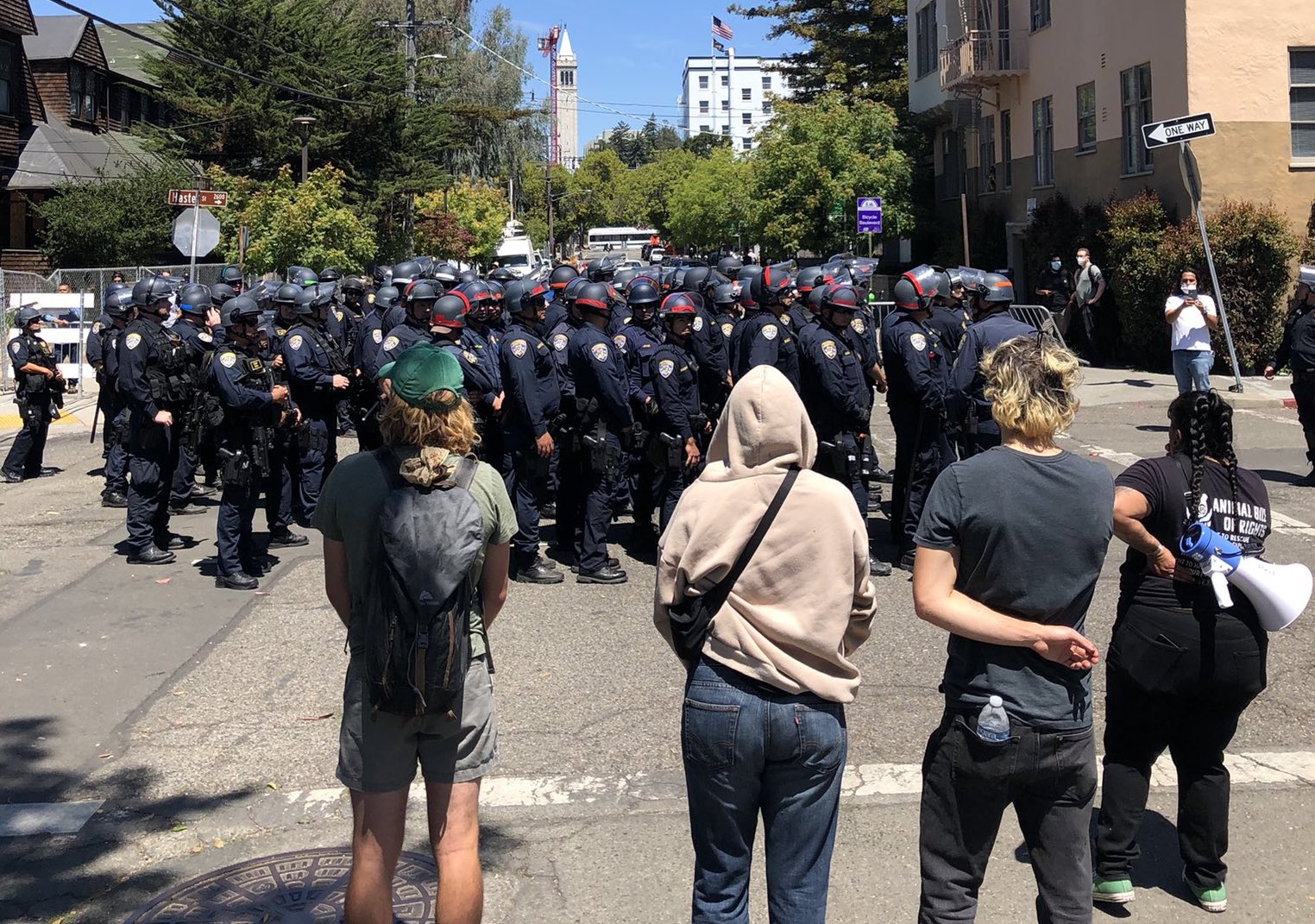 People on the street watch as a large group of police in riot gear stand in a group.