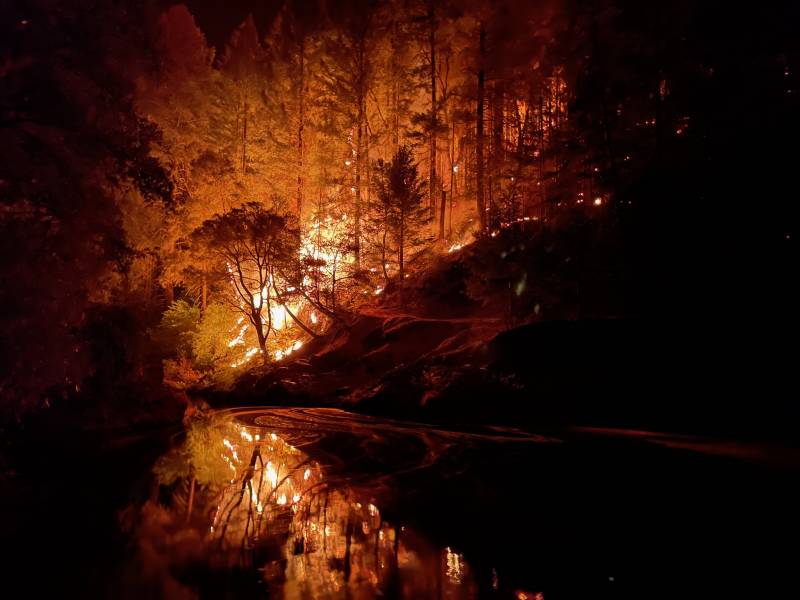 White-yellow flames and an orange sky show through shadows of burnt trees. The flames and colors and reflected in a stream to the lower left of the image. A dark embankment is in the center right of the image.