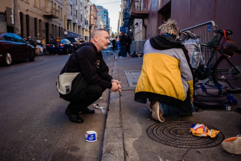 A man crouches down to speak to another person kneeling on the sidewalk in an alley.