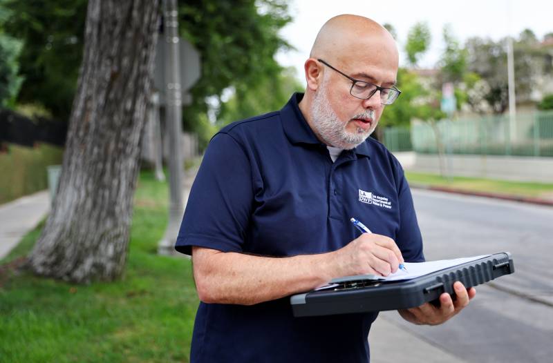 A bald man with light skin and a close-cropped silver beard makes notes on a tablet. He's wearing a blue short-sleeved shirt with Los Angeles Department of Water and Power embroidered in white. Behind the man is blurred green grass and a tree trunk.