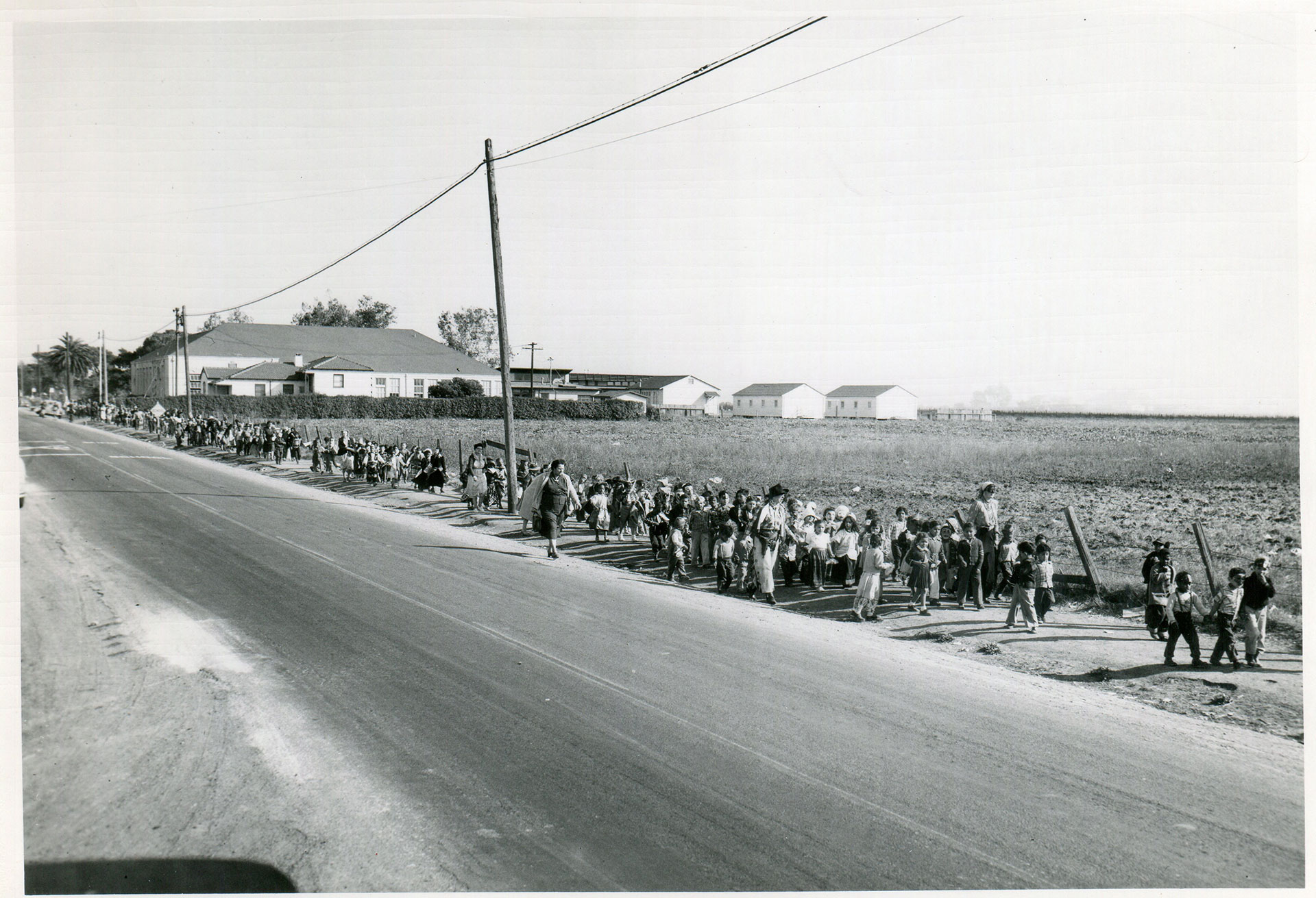 Black and white photo of about fifty children and several adults walking next to a road and a field. The children are dressed up as if for Halloween.