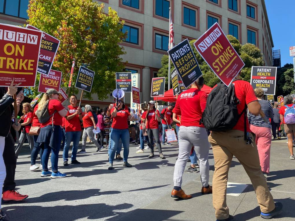 Workers in red shirts holding signs that say 'NUHW on strike' march on a picket line in front of a large building, with one person talking into a bull horn