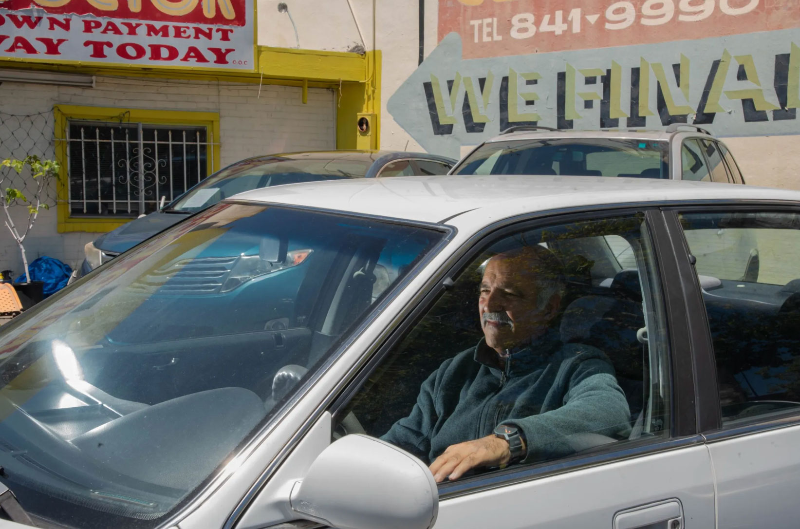 man sits inside car with window down, with car dealership signs in background