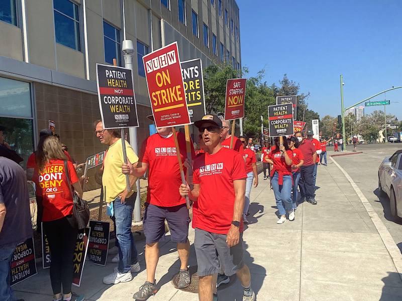 A line of people in red shirts march down a street, carrying signs at a picket line that say 'NUHW on strike.'