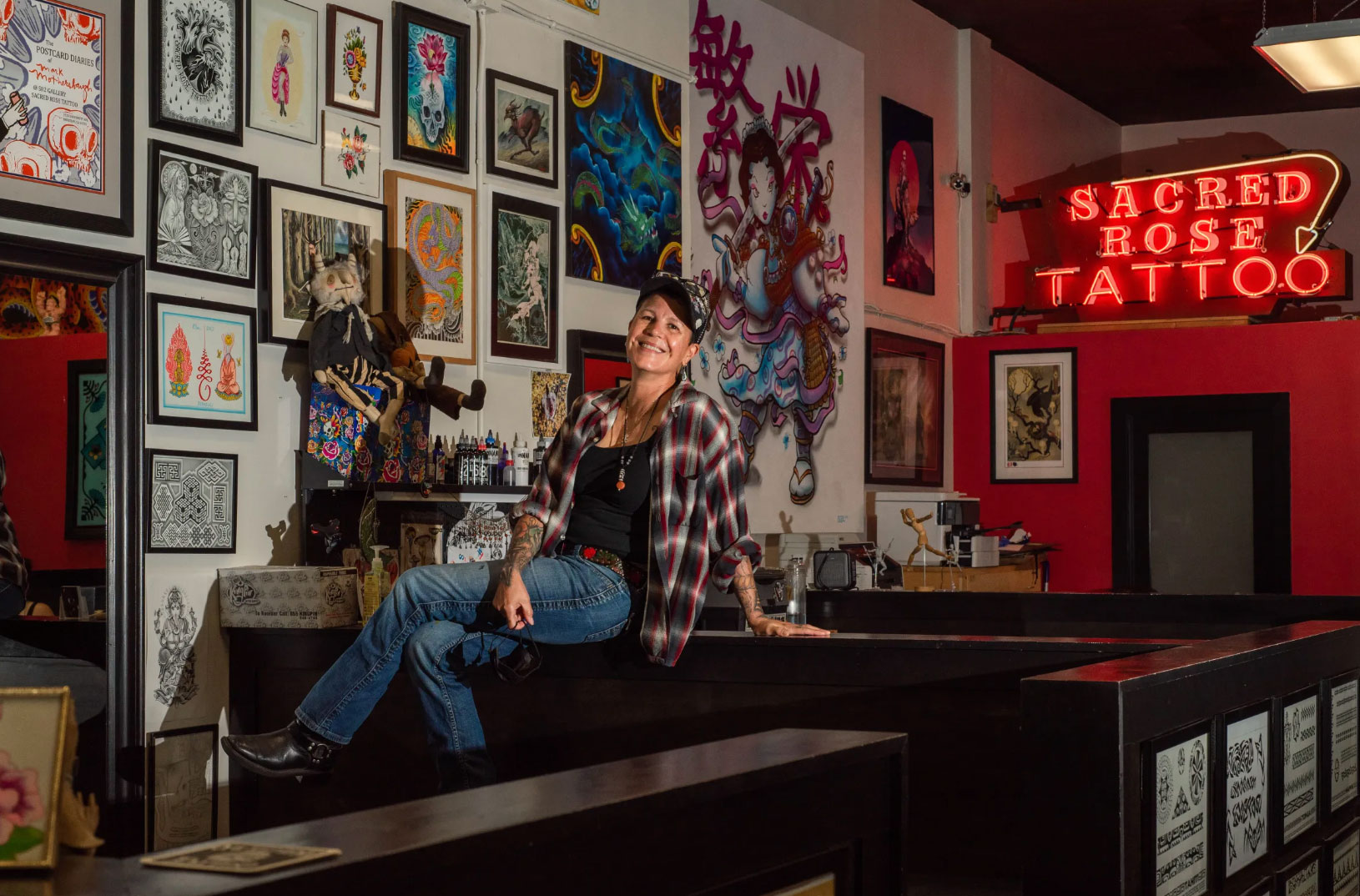 woman wearing jeans and plaid shirt smiles sitting in her tattoo shop, with neon sign in background