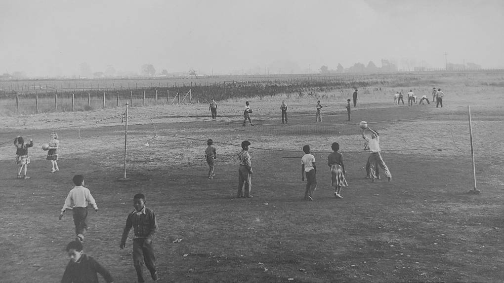 Black and white photograph of kids playing volleyball by an empty field. Some low buildings rise up in the background