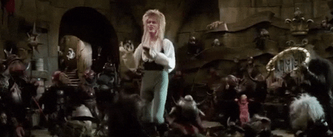 David Bowie dances around with a number of small goblin puppets. He is wearing rather tight grey leggings and a puffy white shirt. 