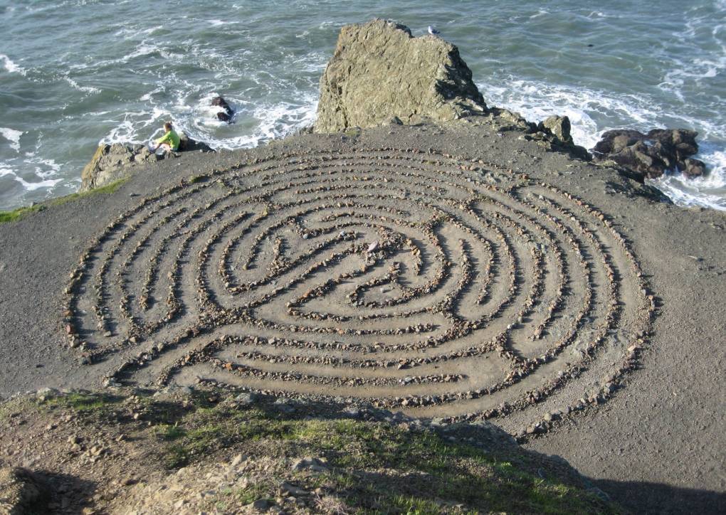 A large Medieval style labyrinth made from stones is laid out on a point of land overlooking a rocky coastline. The ocean is visible just beyond the edge of the labyrinth.