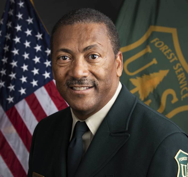 An image of a man with coffee skin and short-cropped hair, wearing a black uniform with a forest service patch on the arm, in front of an American flag and a U.S. Forest Service flag. The first service flag is dark green with an orange logo .