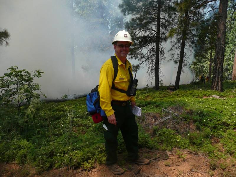 A man with pale skin wearing a white hardhat, yellow shirt and black pants stands on a cleared area of soil and green grasses, with trees and white smoke in the background. He carries a map and wears a pack at the small of his back and a walkie talkie in front.