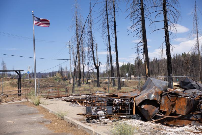 An American flag flies atop a sliver pole at the corner of a cement pad covered by burnt and rusted metal. A row of burnt trees are in the background. 