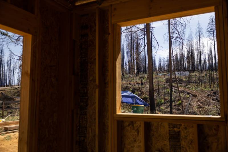 A view through a window from inside a home being built. The view shows a tent in the foreground and a trailer in the background, with burnt trees and patches of green growing from the burnt ground.