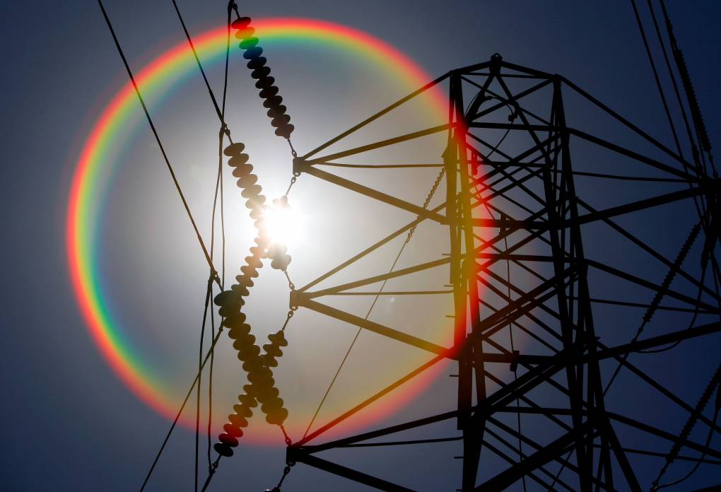 A rainbow around a bright sun shines through an electricity tower and heavy-duty power lines.