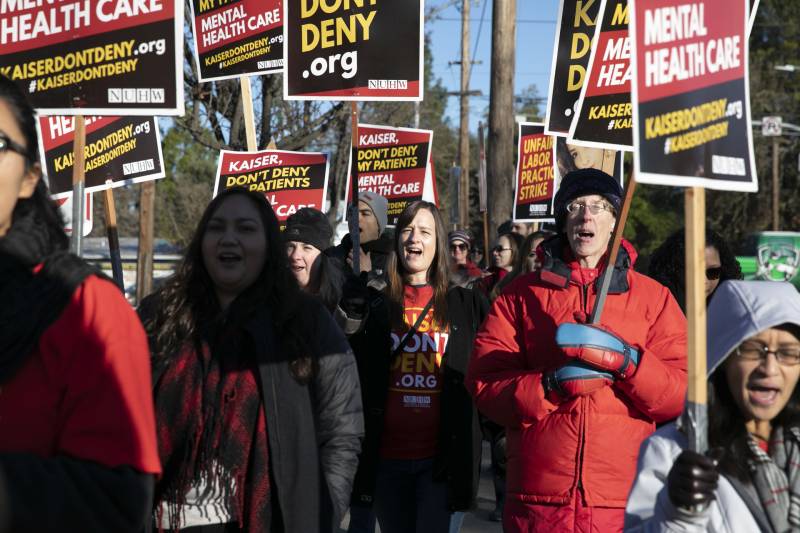 People chanting marcch toward the camera carrying signs reading "Kaiser Don't Deny" in bright yellow lettering on red and black backgrounds. The marchers wear red jackets, and t-shirts bearing the same slogan.