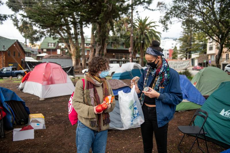 Two people in masks and jackets hold plastic bags and vegetables outside near a tent in a park.