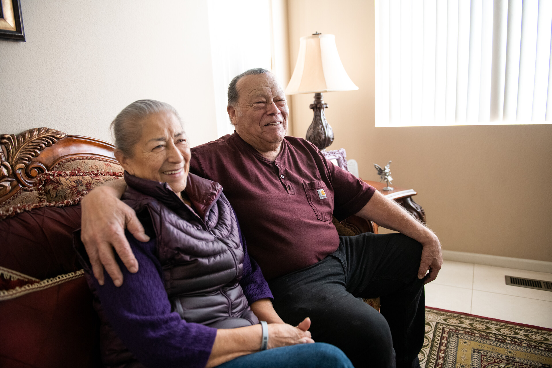 An older Latino couple sits on a couch laughing. The man has his arm around his wife.