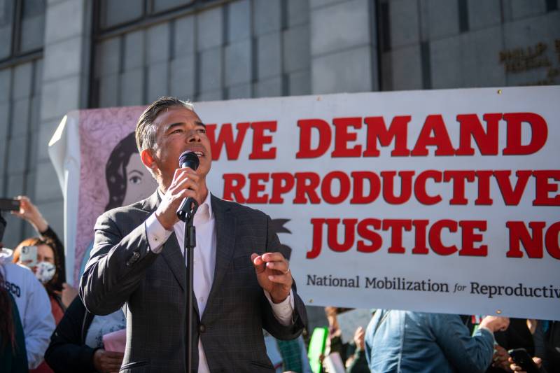 A man in a suit stands in front of a sign saying 'We Demand Reproductive Justice'