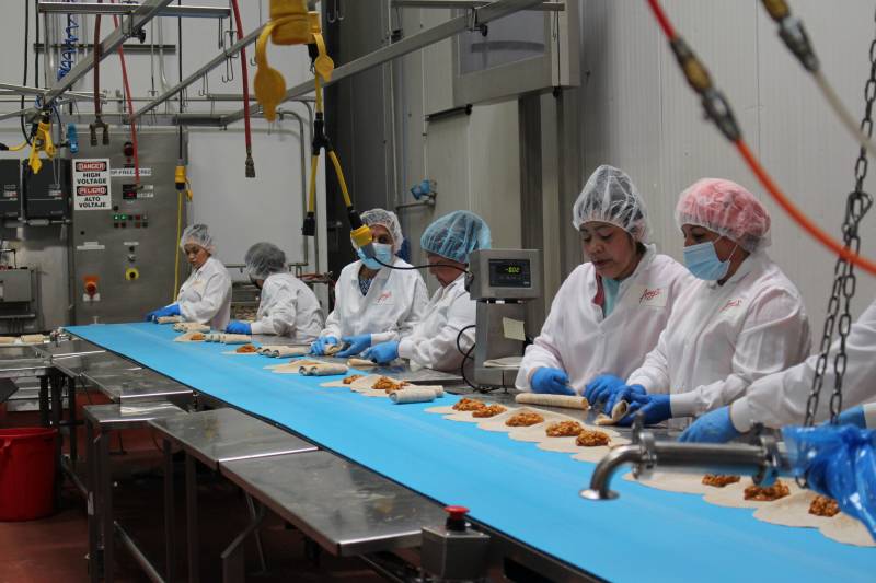 People wearing white coats, hairnets and face masks stand over machines over an assembly line.